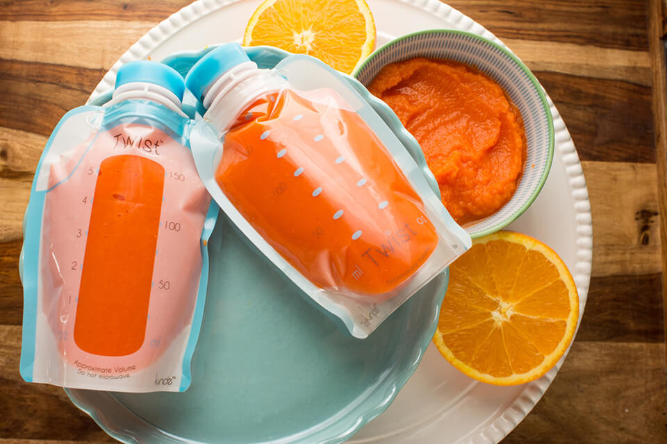 Two Kiinde Storage Bags filled with carrot puree next to a bowl with carrot puree and two orange halves