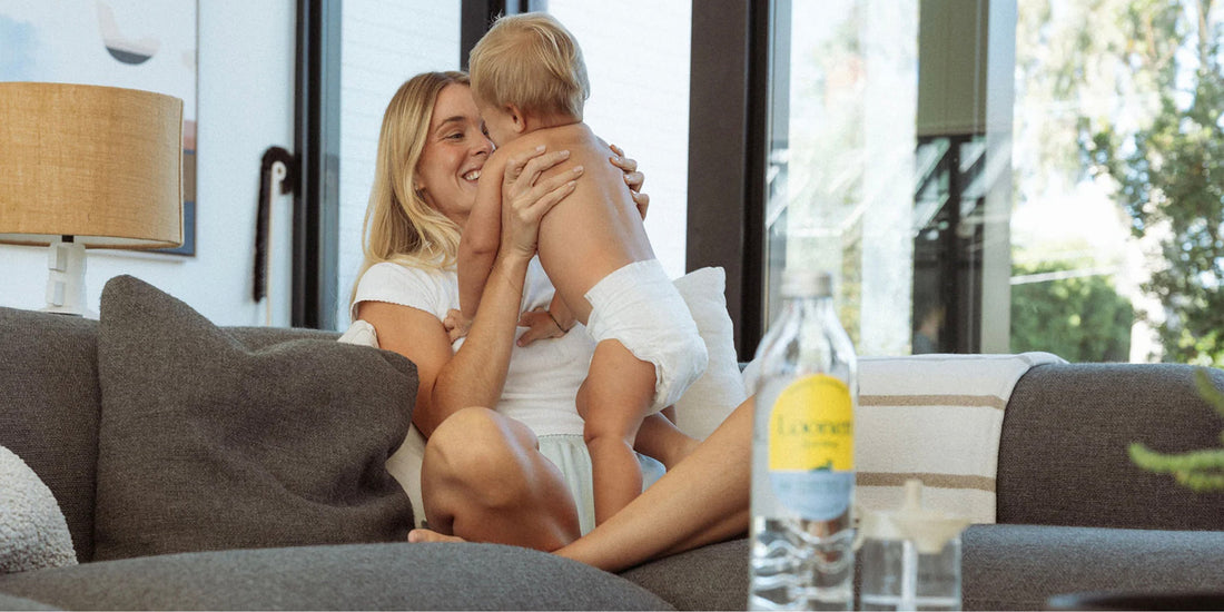 Image of a mom sitting on a couch and holding up her baby with a Loonen glass water bottle on the table in front of her. 