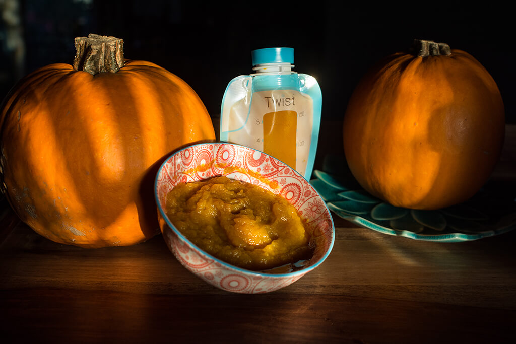 A Kiinde breastmilk storage pouch filled with pumpkin puree behind a bowl of pumpkin puree. There is a pumpkin on either side of the pouch and bowl.
